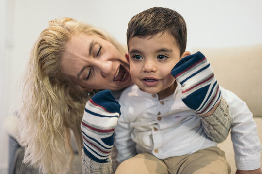 Middle-aged blonde woman looking at and playing puppets with socks in her hands with her young autistic son sitting on her.