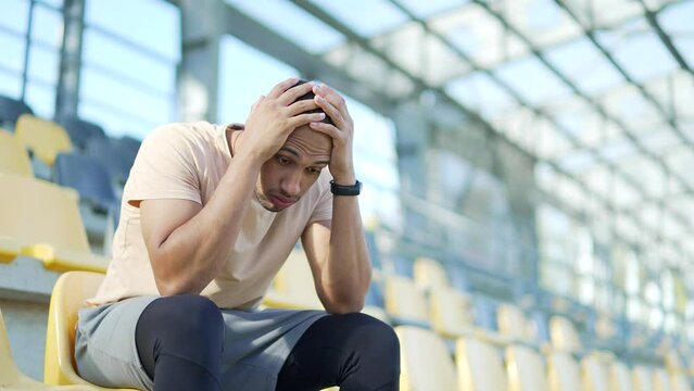 Tired young sportsman in a bad mood sitting in stadium. He looks down at the ground, feeling defeated and frustrated after a tough practice session. He struggles to shake off his negative emotions