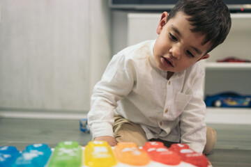 Close-up frontal image of a young autistic boy kneeling on the living room floor looking closely at his musical key toy car. - obrazy, fototapety, plakaty