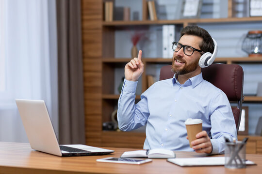 Mature boss investor at workplace inside home office dancing having fun listening to music in headphones, businessman in shirt resting and happy sitting at table using laptop at work.