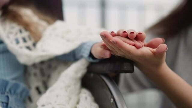 Caring mother strokes hand of little daughter sitting in wheelchair on blurred background. Woman shows care to schoolgirl with muscular dystrophy closeup