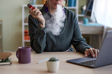 Close-up of of young creative female designer smoking electronic cigarette by workplace while...