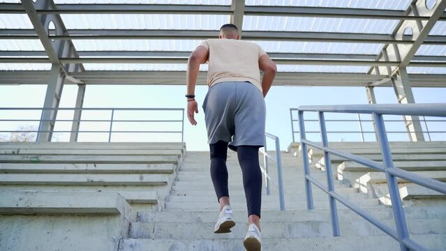 Rear view. Young adult athlete of mixed race running up the stairs doing a workout. A man in a tracksuit trains at an urban stadium lifting on the finish his hands up a triumph gesture. Sport concept
