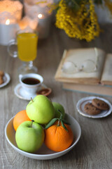 Cup of tea, plates with cookies, glass of orange juice, books, reading glasses, bowl of fruit and candles on the table. Selective focus.