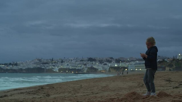 A little girl of five years old dressed in a blue jacket is building something out of sand on the shore of the ocean in the evening. The blue evening light illuminates everything, and in the distance