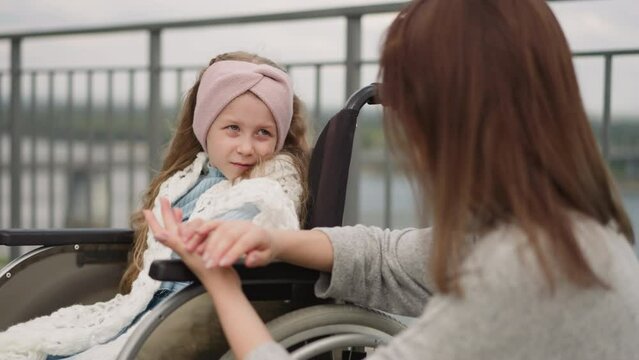 Redhead mother strokes hand of daughter with paralysis expressing love. Cute little girl with headband sits in wheelchair looking at mom against fence closeup