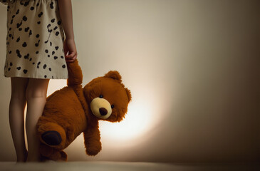 A little girl holds a teddy bear / teddy / stuffed animal in her hand. Background: Neutral Wall. The image symbolises sadness, loneliness. Space for text.