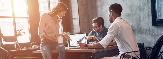 Three confident young people discussing business strategy while pointing laptop in office