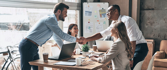 Two young men shaking hands while having meetings with colleagues in the office