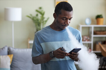 Young African American businessman scrolling in smartphone and smoking electronic cigarette while...