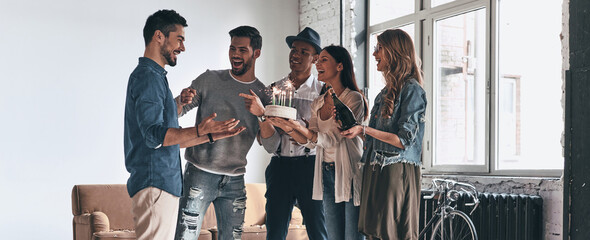 Happy young people holding birthday cake while congratulating their surprised friend
