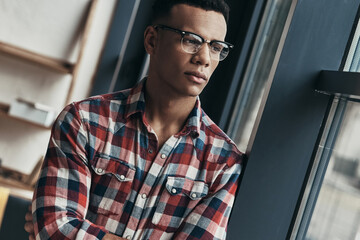 Thoughtful young African man in eyeglasses looking through a window