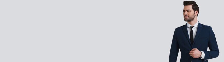 Handsome young man in formal wear adjusting jacket while standing against grey background