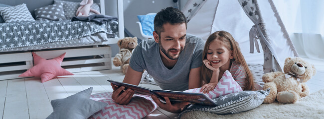 Father reading a book to his little daughter while lying on the floor at home together