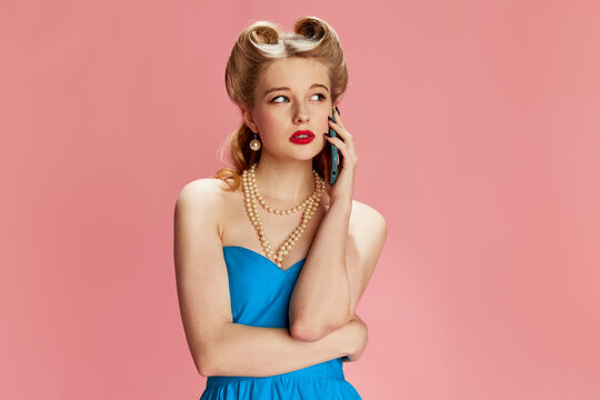 Portrait of beautiful young girl in blue dress and stylish hairstyle posing, talking on phone against pink studio background. Concept of retro fashion, beauty, 50s, 60s. Pin-up style