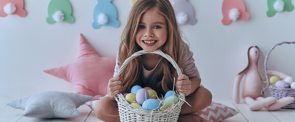 Cute little girl carrying basket with Easter eggs while sitting on the floor with decoration in the background