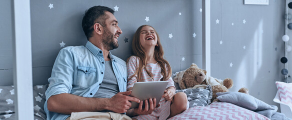 Happy father and his little daughter using digital tablet while sitting on the bed at home together