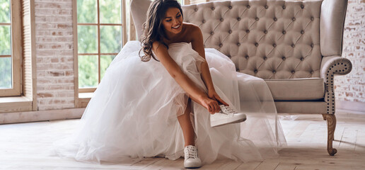 Modern young woman in wedding dress putting on sports shoes and smiling while sitting on the couch - Powered by Adobe