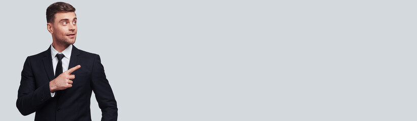 Handsome young man in formal wear pointing copy space while standing against grey background