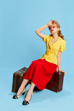 Feeling tired. Beautiful young girl in yellow blouse and red skirt sitting on vintage suitcases against blue studio background. Concept of retro fashion, beauty, travelling, 50s, 60s. Pin-up style