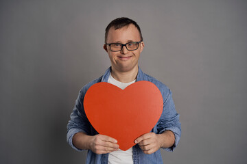 Caucasian man with down syndrome holding big red heart on gray background © gpointstudio