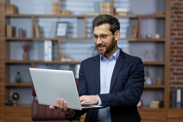 Businessman standing with laptop working inside office, male lawyer mature adult in business suit and beard, holding laptop smiling contentedly.