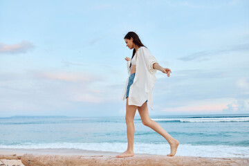 Fototapeta na wymiar Happy tanned woman in white swimsuit shirt and denim shorts walks the beach on a log on the sand by the ocean with wet hair after swimming, sunset light in Bali