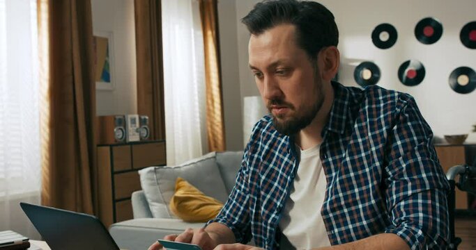 Serious delighed successful young beard man person with disabilities in wheelchair holding debit credit card in hands typing card number on laptop