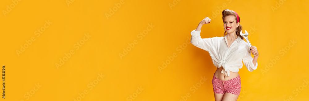 Wall mural Strong hands. Portrait of beautiful young girl in white shirt and pink shorts posing with tool, wrench against yellow studio background. Retro fashion, beauty, 50s, 60s concept. Pin-up style. Banner