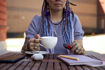 Close-up of young woman with cup of coffee and electronic cigarette sitting by table in outdoor cafe at break and enjoying rest