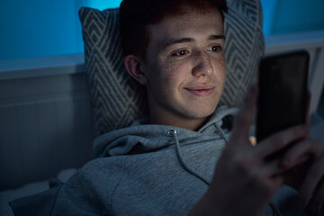 Cheerful caucasian teenage boy using mobile phone while lying in bed at night in his room