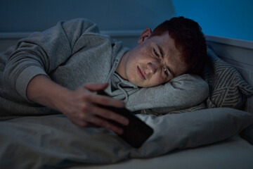 Caucasian teenage boy browsing phone while lying on bed at night