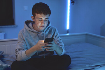 Affectionate caucasian teenage boy using mobile phone while sitting at night in his room