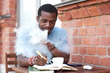 Young African American businessman smoking electronic cigarette and making notes in notebook while...
