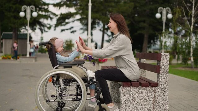 Mother and daughter with spinal paralysis play patty in city park. Young woman sits on bench entertaining little girl in wheelchair on spring day