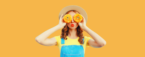 Summer portrait of happy young woman covering her eyes with flowers as binoculars looking for something wearing round straw hat, jumpsuit on orange background