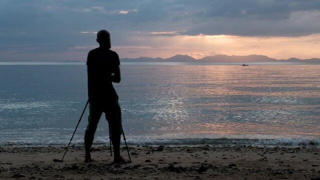 Tripod shot of a tourist (photographer) taking a sunset on a camera mounted on a tripod on the beach. Klong Muang Beach, Krabi Province, Thailand.