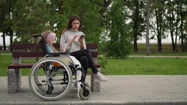 Woman checks email while little girl in wheelchair watching videos on smartphone. Focused mother and daughter use gadgets after walk in city park