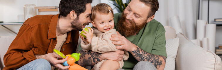 Positive same sex parents looking at baby daughter with apple at home, banner.