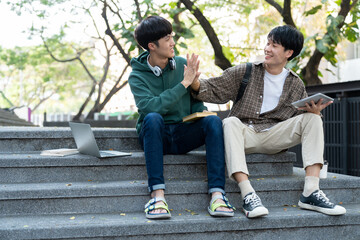 Two Asian male students sitting on the stairs of the university enjoy chatting after school using smartphones and tablets to find information together.