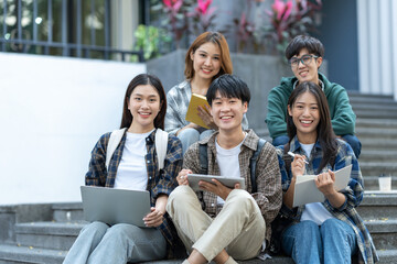 Group of happy young Asian students in casual clothes sitting on the front steps of a university building looking for educational information. teamwork classmate best friend.