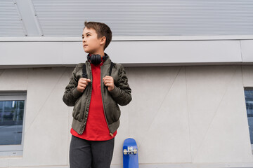preteen boy in bomber jacket and wireless headphones standing with backpack near penny board.