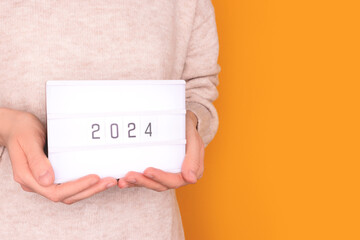 Female hands hold lightbox with 2024 numbers in front of yellow background. New Year concept with...