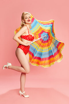 Summer vacation. Portrait of beautiful young woman in swimming suit posing against pink studio background with sun umbrella. Concept of retro fashion, beauty, 50s, 60s. Pin-up style