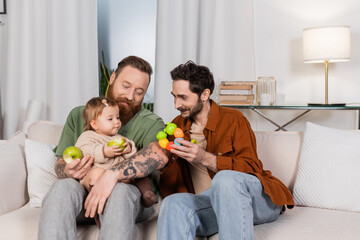Homosexual couple holding toy near baby daughter with fresh apple on couch at home.