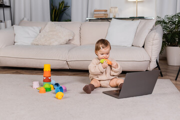 Baby girl holding toy near laptop on floor at home.