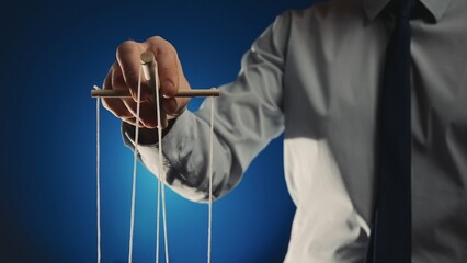 A businessman in a gray shirt and black tie controls a puppet with a wooden manipulator and strings. Hand handling at puppet by pulling strings to make the character move. Blue background. Close up.