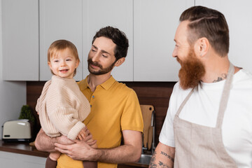 Fototapeta premium Gay parent holding smiling toddler daughter near husband in kitchen.