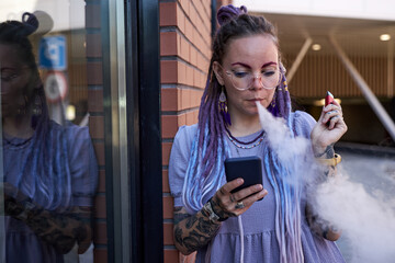 Young woman with dreadlocks looking at smartphone screen and smoking electronic cigarette while...