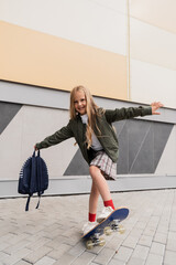 full length of happy preteen girl in stylish bomber jacket holding backpack while riding penny...